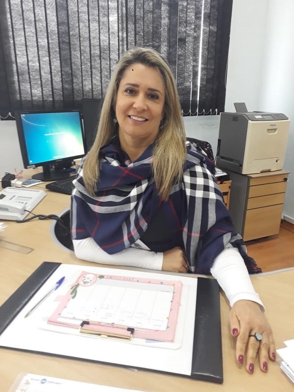 Foto mostra mulher branca, loira, sorrindo, sentada em mesa comercial, na mesa há computador e ao fundo persiana fechada de cor cinza. A mulher apresentada na imagem é a Subprefeita de Itaquera. 